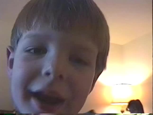 Young boy films himself in a cozy living room while a woman sits on the couch behind him
