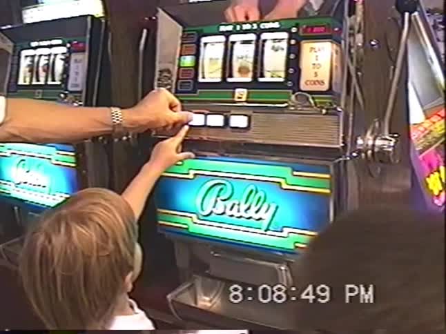 Vintage home video shows a young boy getting help from an adult to play a classic Bally slot machine in a casino