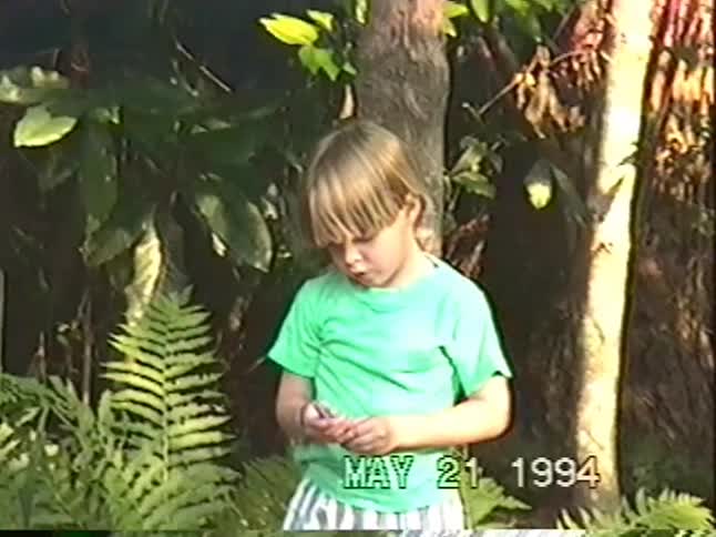 A little boy in a green shirt animatedly talks to the camera while playing outside in a wooded yard in 1994