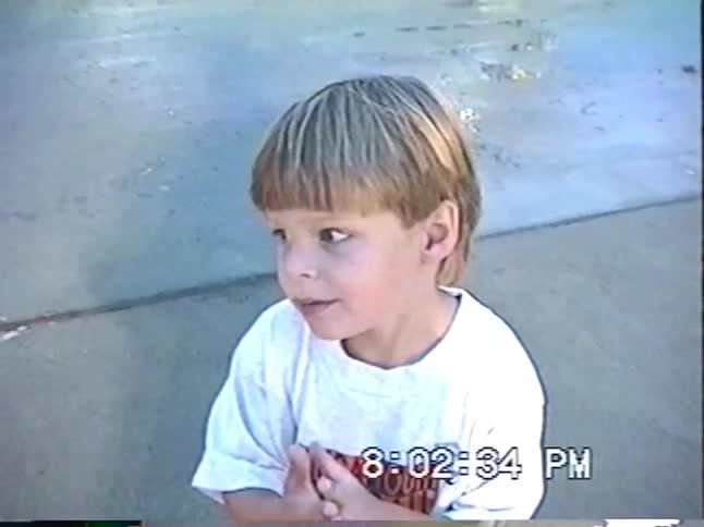 Vintage VHS footage shows two young boys excitedly pointing and talking at a 'Kiddieland' amusement park in the evening
