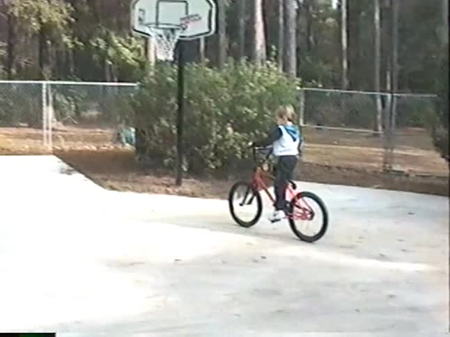 A proud young boy smiles for the camera before happily riding his red bicycle with a front basket around the driveway