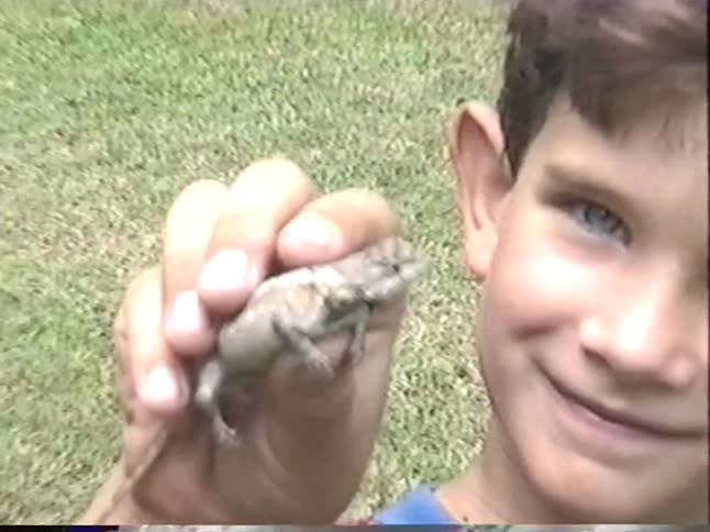 A smiling young boy with bright blue eyes holds a small lizard up to a vintage home video camera during a family gathering