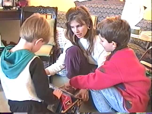 A mother and her two young sons look at a new toy together in the living room in this vintage home video