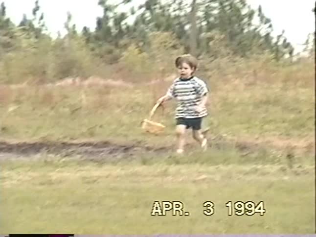 A vintage VHS recording of a young boy happily running with his basket during an Easter egg hunt in a field in 1994