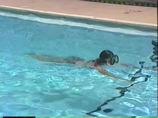 Vintage home video footage shows a young woman in a snorkel mask leisurely swimming in an outdoor pool on a bright, sunny day