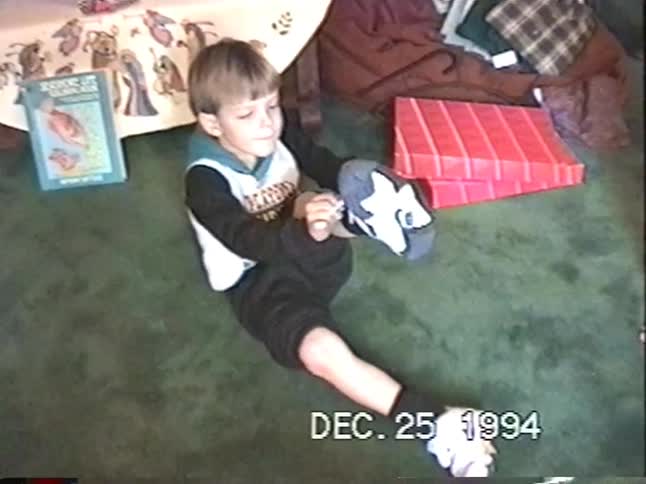 Vintage VHS footage from Christmas 1994 shows children opening presents, with one boy excited to try on his new pair of sneakers