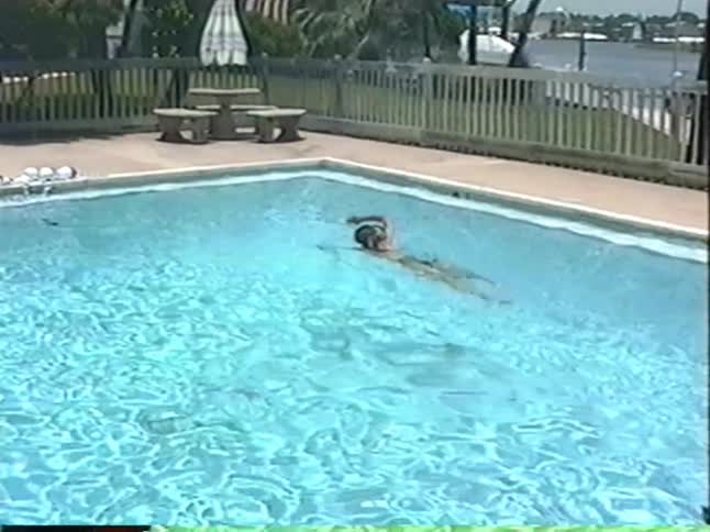 A woman wearing a snorkel mask swims laps in an outdoor pool in grainy, vintage home video footage