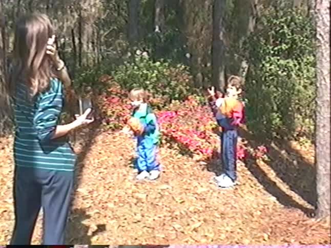 Vintage home movie of a mother taking a picture of her two young sons posing with baseball gloves in front of a flowering bush