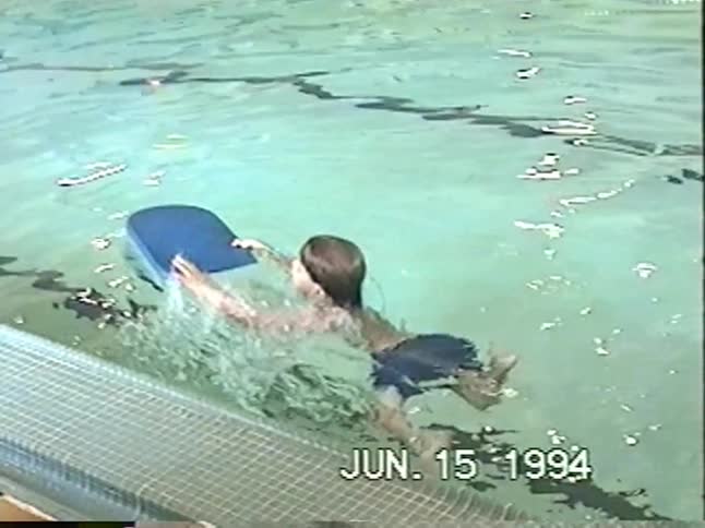 A vintage home video from 1994 shows a young boy learning to kick and swim with a blue kickboard in a swimming pool
