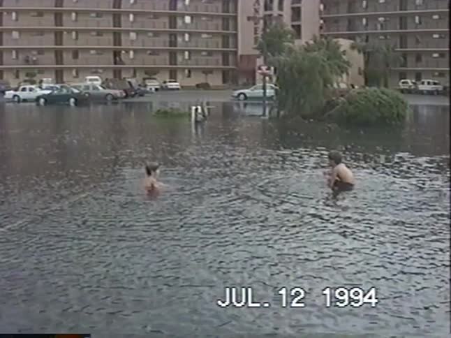 Vintage VHS footage shows two young boys joyfully playing and splashing in a flooded apartment complex parking lot in 1994