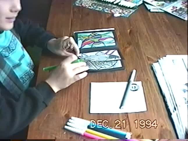 Vintage VHS footage from 1994 captures a young boy intensely focused on his stained glass coloring book at a wooden table before panning to a table of wrapped Christmas gifts