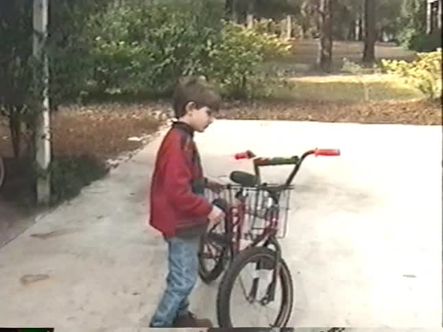 A nostalgic home video from the 1990s shows a mother and her two young sons with their bicycles in the driveway