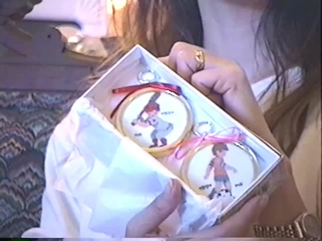 A mother opens a thoughtful Christmas gift revealing two handmade embroidered sports-themed ornaments in a vintage home video