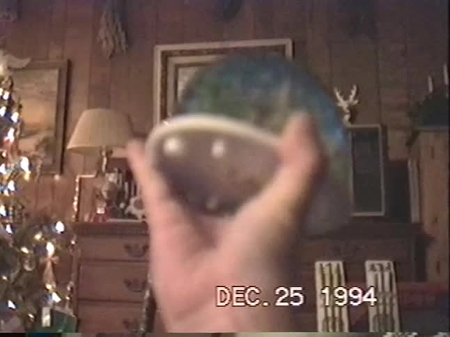 A nostalgic home video from 1994 shows a young boy excitedly opening a snow globe as a Christmas present while his brother watches by the tree