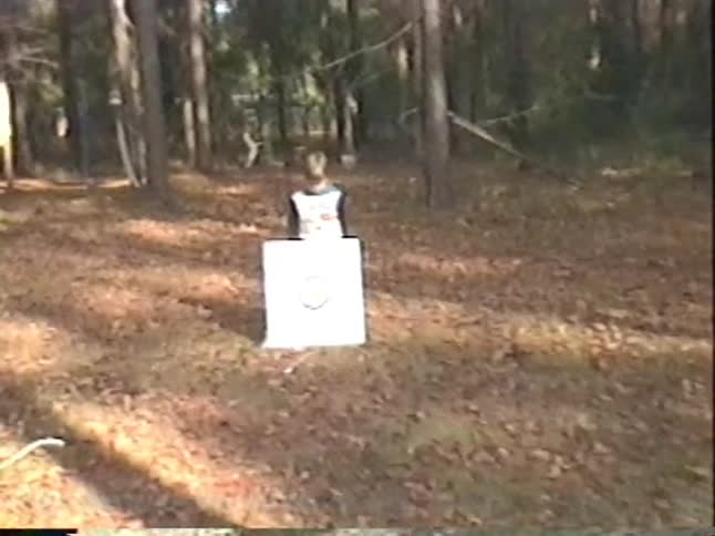 Vintage home movie footage captures a father teaching his two young sons how to shoot a bow and arrow at a target in their wooded backyard
