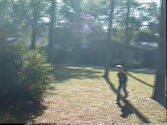 Vintage home video footage shows a young blond boy running through a sunlit, wooded backyard