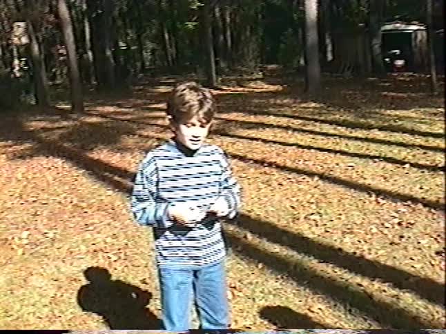 Vintage home video footage captures a young boy in a striped shirt playing with a yoyo in a sun-dappled, wooded backyard