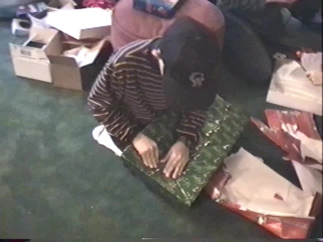 A young boy in a baseball cap excitedly unwraps a large Christmas present in a living room
