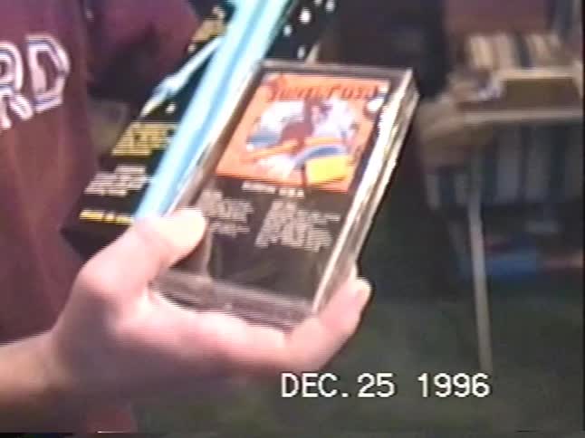 A person proudly displays their new "Surfin' U.S.A." music cassette tape in a close-up from a 1996 home video