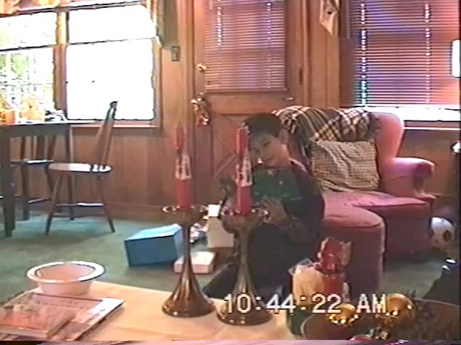 Vintage VHS footage shows a young boy in a baseball cap unwrapping a Christmas present in a cozy living room
