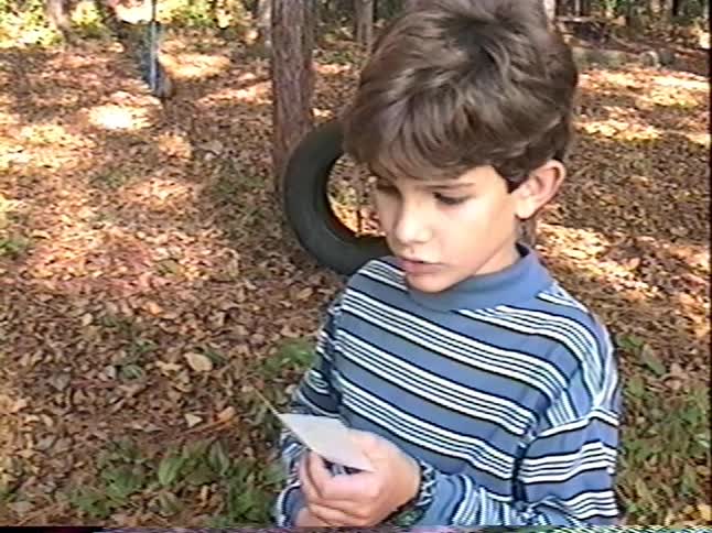 A young boy plays with a paper fortune teller in his wooded backyard in this nostalgic home video