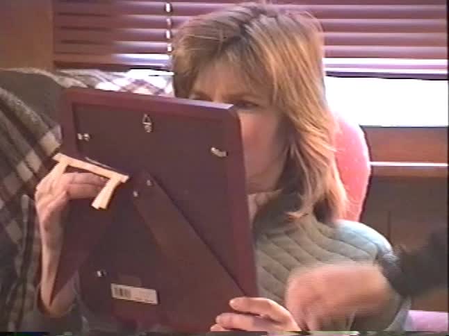 A vintage VHS recording shows a woman unwrapping a picture frame and a small Harvard pennant while sitting in a cozy armchair