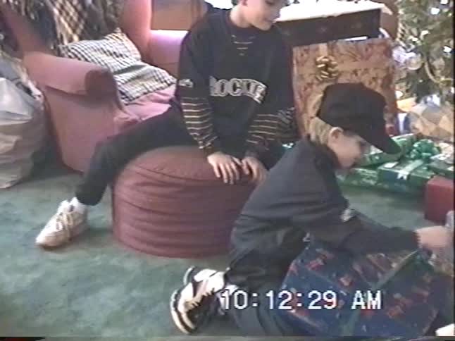 Vintage VHS footage shows two brothers on Christmas morning, with one excitedly opening a large present by the tree