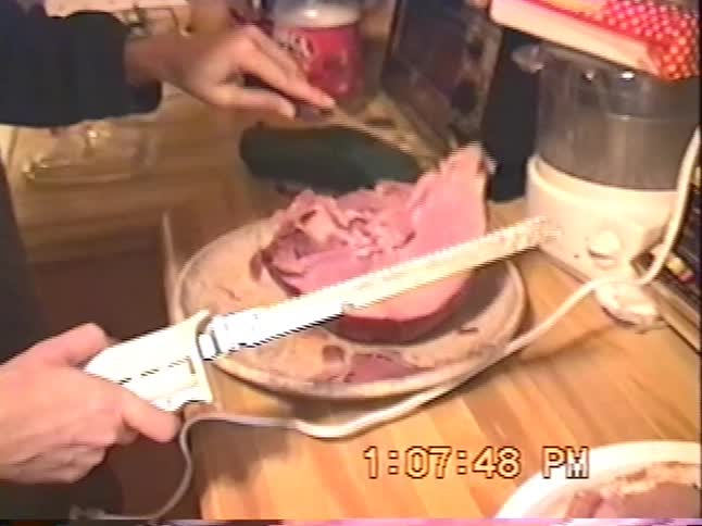A person uses an electric knife to slice a large baked ham on a wooden kitchen counter in this authentic VHS home video