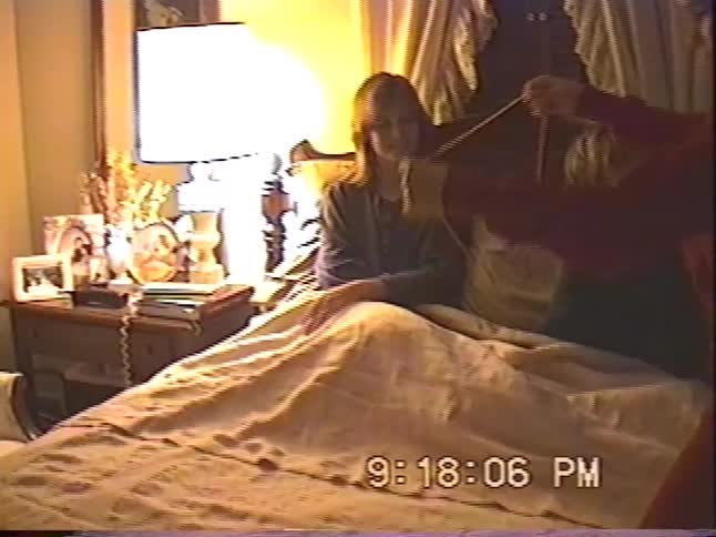 A young boy proudly displays his Cat's Cradle string game skills for the camera in a warmly lit bedroom during a family home video session
