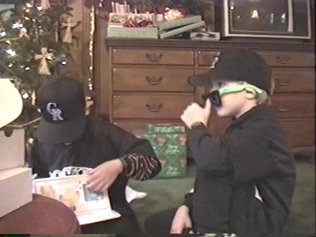 Vintage VHS footage shows two brothers in matching baseball jerseys opening Christmas presents, with one dancing excitedly in new sunglasses