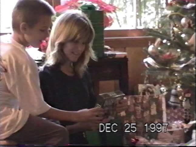 Vintage VHS footage shows siblings opening a present together in front of the Christmas tree in 1997