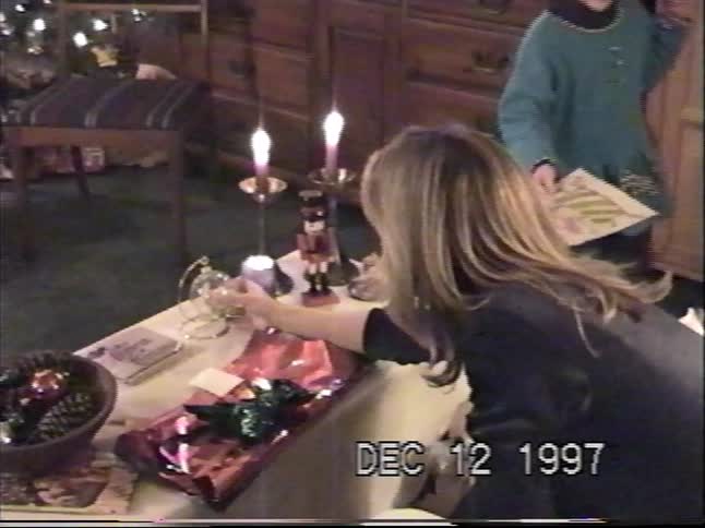 A nostalgic 1997 home video shows a woman unwrapping a glass ornament during a cozy, candlelit Christmas gift exchange