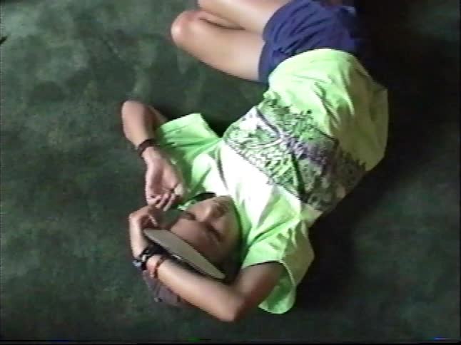 A teenager in a neon shirt and backwards cap lazily rolls across the green carpet in vintage home movie footage