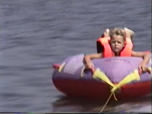Vintage VHS footage shows a young blonde girl in a life jacket having fun while tubing on a lake