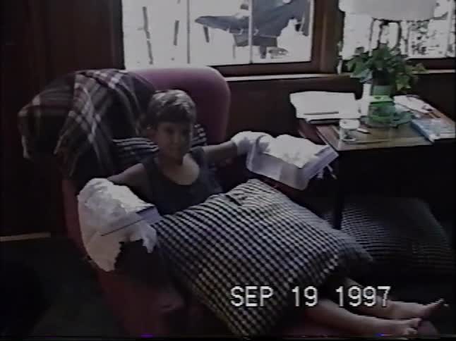 A young boy with both hands in large bandages sits comfortably in an armchair and smiles in a 1997 home video
