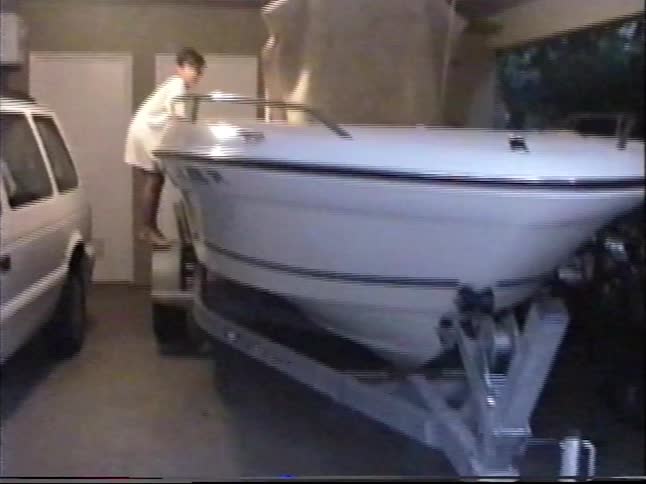 A young boy climbs on a motorboat parked on a trailer inside a home garage in this vintage home video