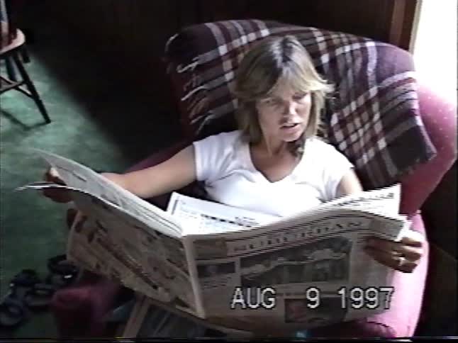 Vintage VHS footage from 1997 shows a woman casually reading a newspaper in a cozy armchair
