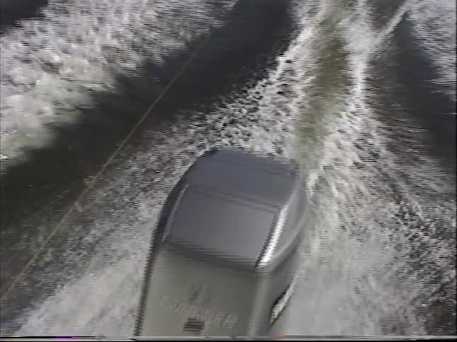 Vintage VHS footage from the back of a speeding motorboat shows a person enjoying a fun ride on an inflatable tube across a lake