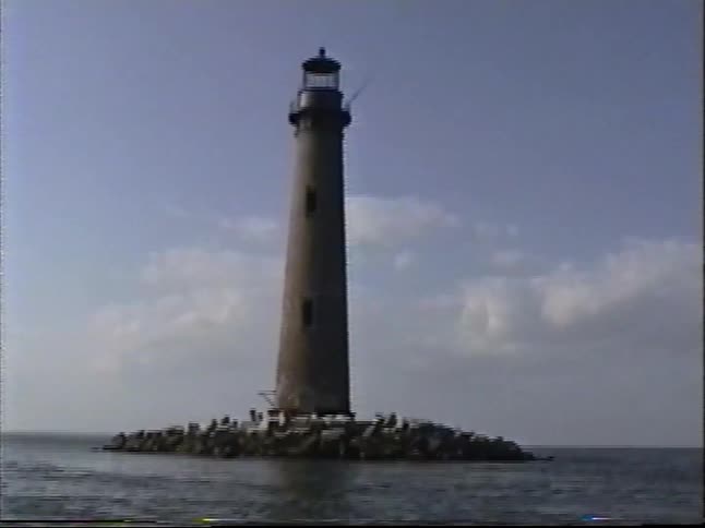 A vintage, handheld shot shows a tall, solitary lighthouse built on a small rocky island in the calm ocean