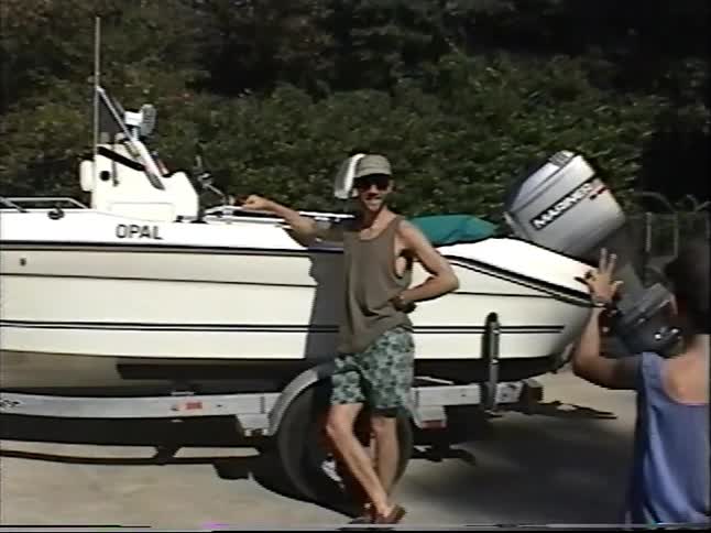 Vintage VHS footage shows a proud father posing next to his motorboat on a trailer as his family films him on a sunny day