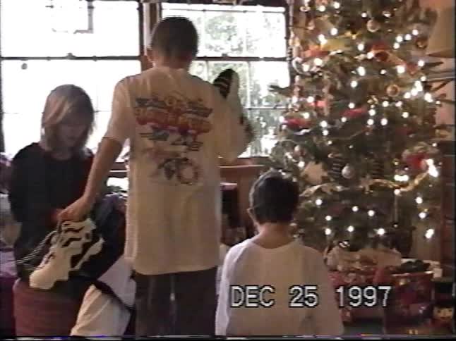 A family joyfully unwraps Christmas presents next to their decorated tree in a classic 1997 home video