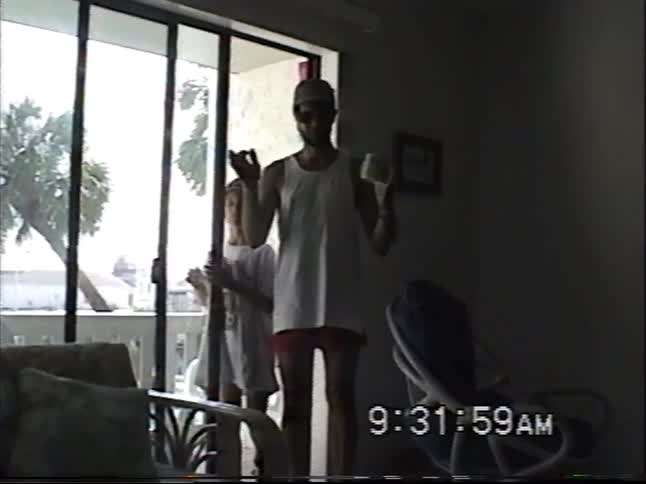 A father and son walk in from the balcony of their vacation home and playfully pose for the camera in this vintage home video