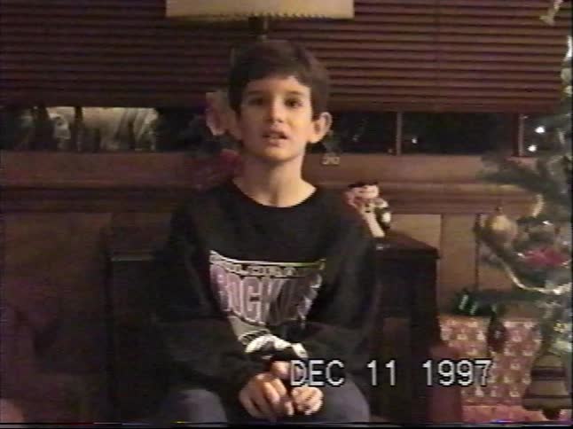 A young boy sits in front of a decorated Christmas tree and talks to the camera in a nostalgic home video from 1997
