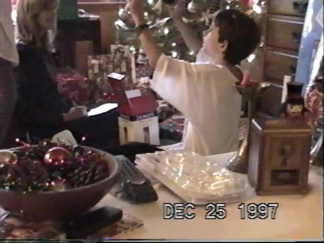 A young boy opens Christmas presents with his family in front of a decorated tree in this vintage home video from 1997