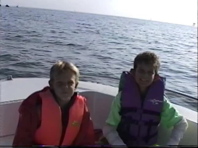Two young boys in life jackets enjoy a boat ride on the ocean, passing by a tall lighthouse