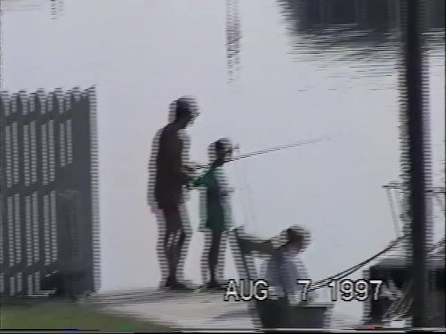 Vintage 1990s home movie footage shows a family fishing off a boat dock behind a swimming pool on a sunny day