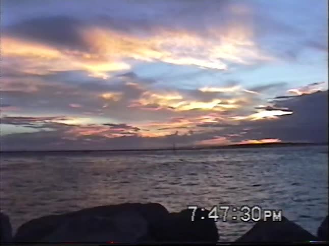 A colorful sunset over the ocean from a rocky jetty pans to reveal a man enjoying the view from a white sand beach
