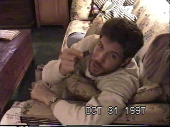 A man lies on a couch talking to the camera and eating a piece of candy in a vintage home video from Halloween 1997