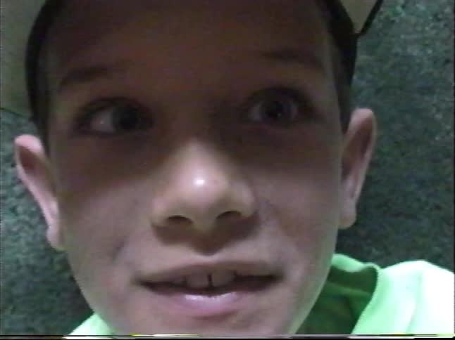 A close-up of a young boy wearing a baseball cap and a green t-shirt as he talks to the camera in a vintage home video