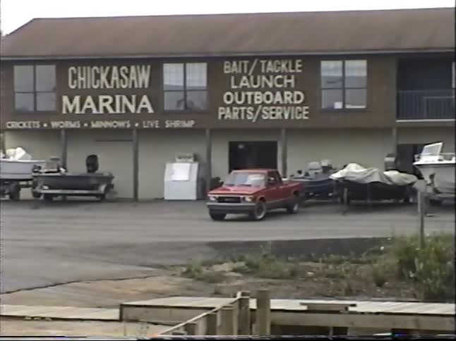 A slow zoom and pan shot of the Chickasaw Marina with boats parked outside and docks on the water, captured on vintage VHS tape